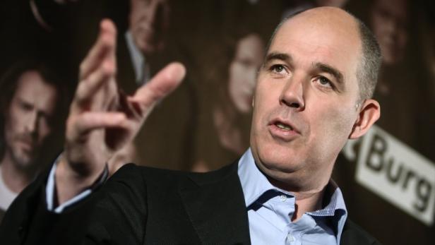 Ein Mann in Anzug gestikuliert vor einem unscharfen Hintergrund.