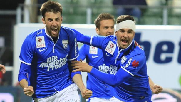 Zwei jubelnde Fußballspieler in blauen Trikots, einer mit Kopfverband.