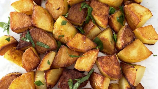 Gebratene Kartoffelstücke, garniert mit frischen Kräutern, liegen auf einem weißen Untergrund.