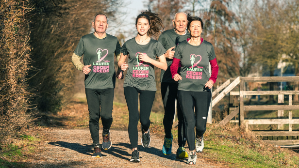 Vier Personen laufen einen Waldweg entlang, sie tragen T-Shirts mit der Aufschrift "Laufen gegen Krebs"