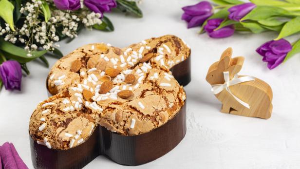 Eine Colomba Pasquale, ein traditioneller italienischer Osterkuchen, mit Mandeln und Zuckerstreuseln, neben einem Holzhasen und lila Tulpen.