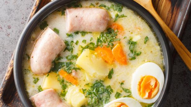 Polnische Suppe Zurek mit Weißwurst, Eiern, Kartoffeln und Karotten in einer schwarzen Schüssel, danaben ein Holzlöffel und Brot