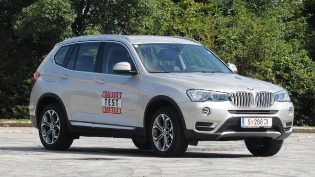 Ein silberner BMW X3 steht vor einer grünen Baumreihe.