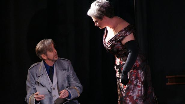 Eine Frau im Abendkleid steht vor einem Mann im Militärmantel auf einer Theaterbühne.