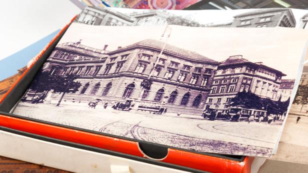 Eine alte Schwarzweißfotografie eines großen Gebäudes mit Straßenbahnen davor in einer Schachtel.