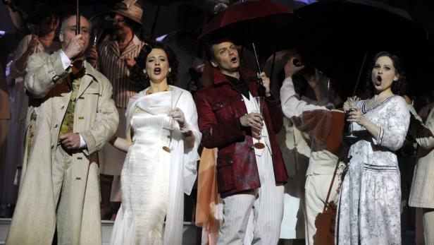 Eine Opernszene mit Sängern, die unter Regenschirmen stehen und singen.