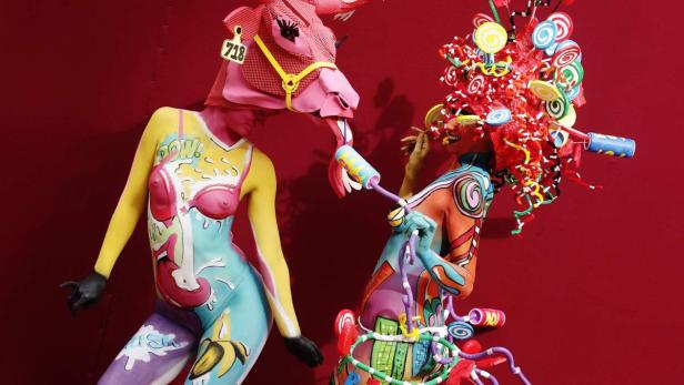 Zwei Frauen mit Körperbemalung und extravaganten Kopfbedeckungen posieren vor einem roten Hintergrund.