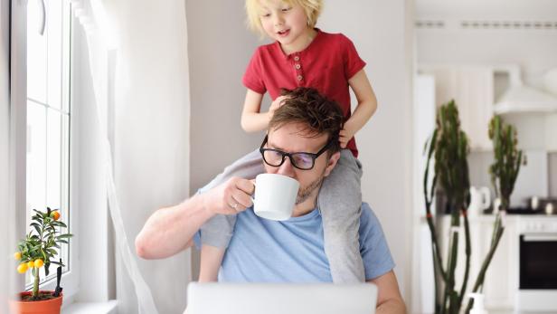 Ein Vater sitzt kaffeetrinkend vor dem Laptop und trägt sein Kind auf den Schultern.