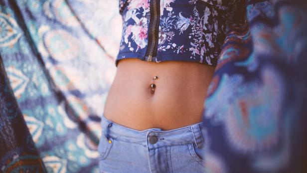 Eine Frau mit einem Bauchnabelpiercing trägt ein kurzes, geblümtes Oberteil und Jeansshorts.