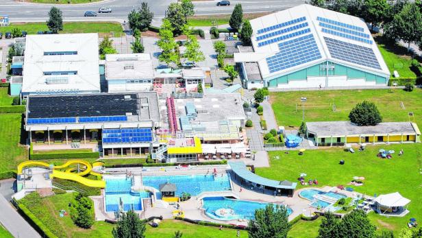 Luftaufnahme eines Schwimmbads mit mehreren Becken, einer gelben Rutsche und Liegewiesen.