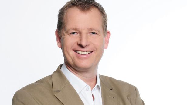 Ein lächelnder Mann in einem hellbraunen Anzug vor einem weißen Hintergrund.