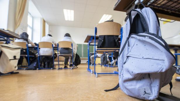 Ein hellgrauer Rucksack steht im Vordergrund eines Klassenzimmers.