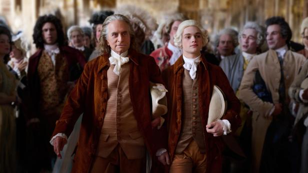 Benjamin Franklin, gespielt von Michael Douglas, geht mit seinem Enkel Temple, gespielt von Noah Jupe, durch eine Menge französischer Männer. Sie tragen braune Anzüge, Temple auch eine weiße Perücke.