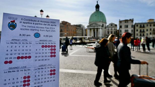 Ein Schild auf einem venezianischen Platz informiert über das Eintrittsgeld ab 25. April. Im Hintergrund gehen Touristen mit Koffern