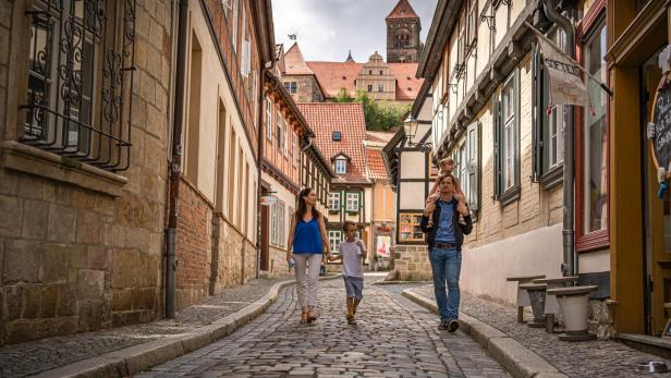 Familie mit Kindern spaziert in mittelalterlicher Stadt in einer Gasse mit Kopfsteinpflaster, im Hintergrund sieht man eine Burg.