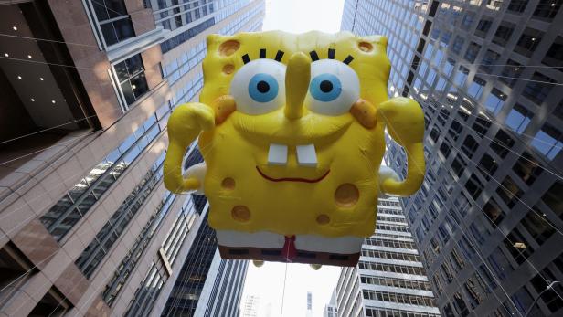 Ein riesiger Ballon, der die Cartoonfigur SpongeBob - ein gelber Schwamm mit Hose an - darstellt, fliegt zwischen Hochhäusern durch
