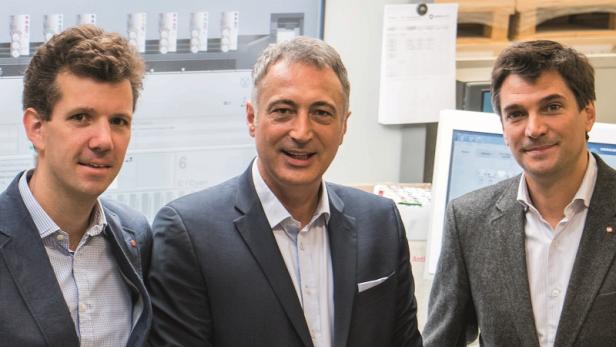 Drei Männer in Anzügen stehen vor einem Bildschirm mit technischen Daten.