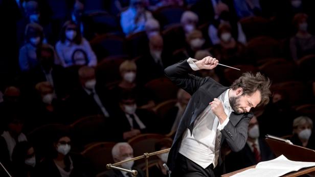 Ein Dirigent leitet ein Orchester vor einem Publikum, das Masken trägt.