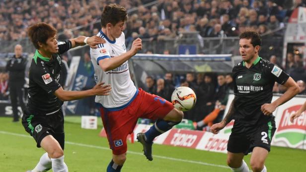 Ein Fußballspieler des Hamburger SV schießt den Ball, während zwei Spieler von Hannover 96 zusehen.