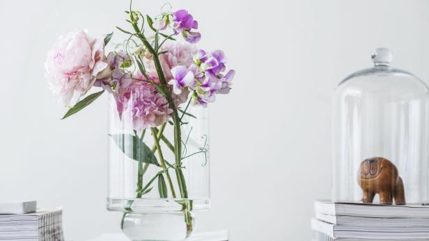 Ein Blumenstrauß mit rosa Pfingstrosen und lila Wicken steht in einer Glasvase auf einem Bücherstapel.