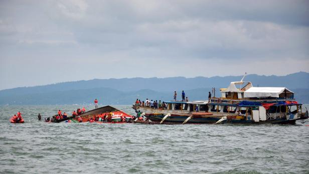 Ein Schiff ist gesunken, während ein anderes voller Menschen daneben liegt; Rettungskräfte sind im Einsatz.