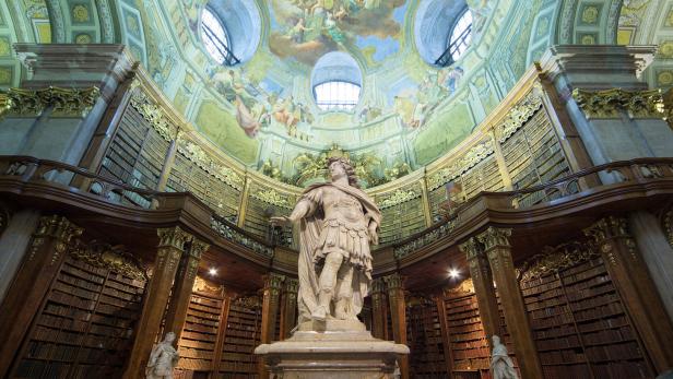 Prunkvoller Bibliothekssaal mit einer großen Statue im Zentrum, umgeben von hohen Bücherregalen und einer reich verzierten Kuppeldecke.
