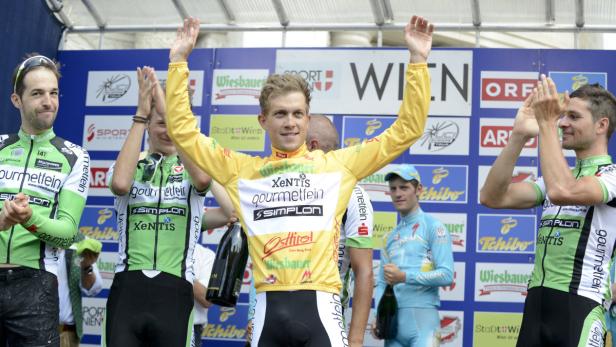 Radrennfahrer auf dem Podium mit erhobenen Armen, umgeben von applaudierenden Teammitgliedern.
