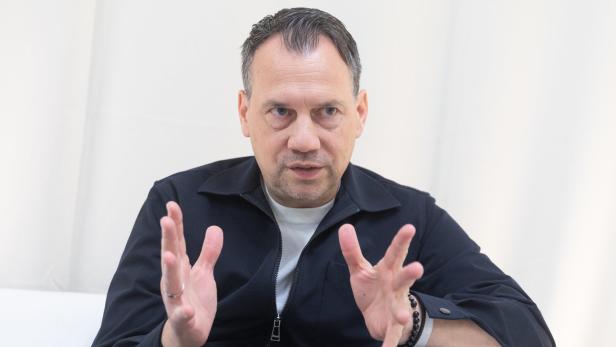 Sebastian Fitzek in dunkler Weste vor weißem Hintergrund gestikuliert und spricht.