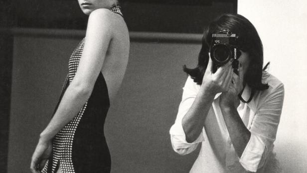 June Newton in weißer Bluse und weißer Hose fotografiert ein Model in einer Abendrobe. Die Kamera verdeckt das Gesicht der Künstlerin, das Model blickt ernst