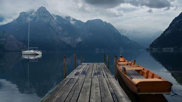Ein See mit einem Segelboot und einem Ruderboot an einem Holzsteg vor einer Bergkulisse.