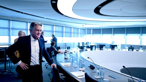 Ein Mann im Anzug, es ist Peter Westenthaler, steht in einem Konferenzraum mit ovalem Tisch und Mikrofonen.