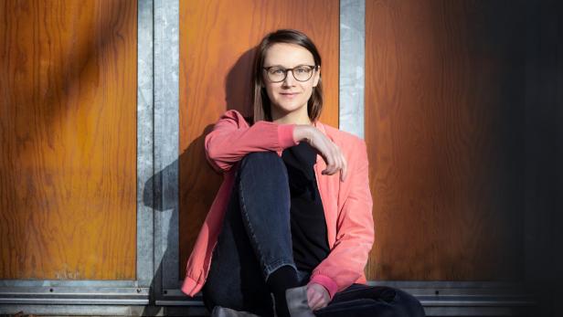 Autron Johanna Sebauer sitzt mit einer roten Jacke vor einer Holzwand