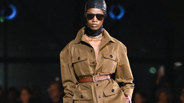 Eine Frau in einem braunen Hosenanzug mit Lederkappe und Sonnenbrille auf dem Laufsteg.