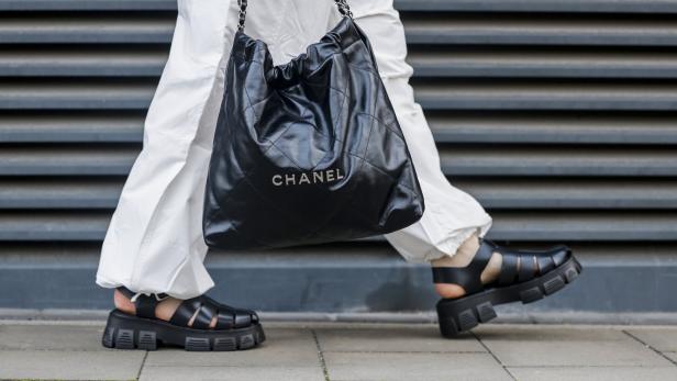 Eine Person mit weißen Hosen und schwarzen Sandalen trägt eine schwarze Chanel-Tasche.