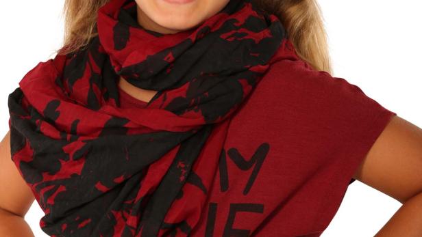 Ein junges Mädchen mit Zöpfen und einem roten T-Shirt mit einem schwarzen und roten Schal lächelt.