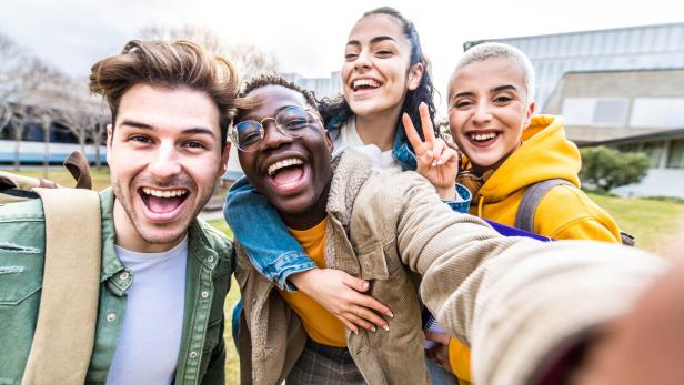Vier junge Leute machen ein Selfie und lächeln in die Kamera, dabei nimmt ein junger Mann eine junge Frag Huckepack