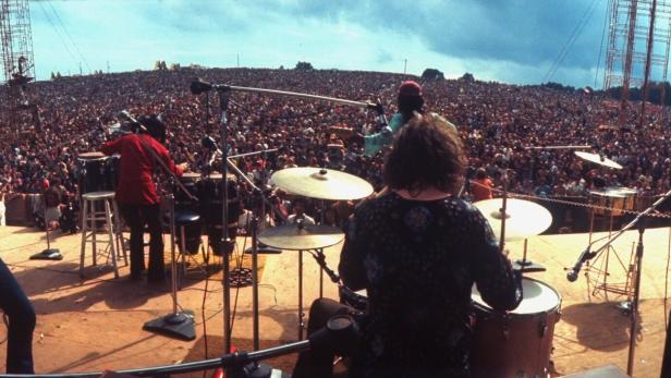 Eine Band auf der Bühne beim Woodstock Festival 1969. Von der Bühne blicken die Musiker inmitten eine Menschenmasse