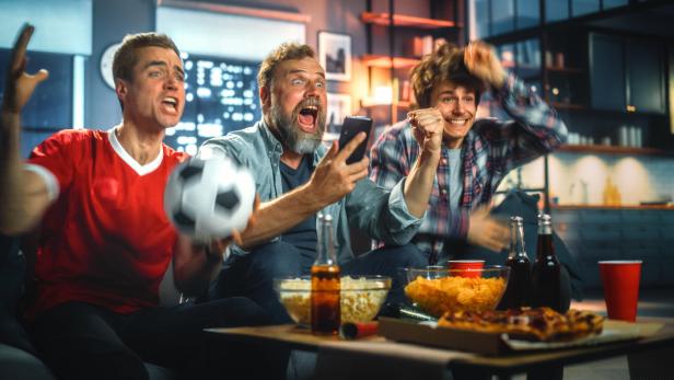 Drei Männer jubeln begeistert beim gemeinsamen Fußballschauen auf dem Sofa mit Snacks und Getränken.