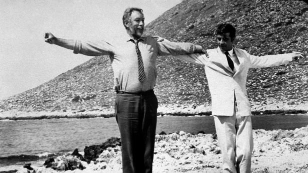 Zwei Männer, möglicherweise Darsteller des Films „Alexis Sorbas“, stehen am Strand mit ausgestreckten Armen.