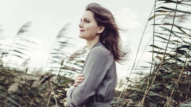 Frau im grauen Kleid steht zwischen hohem Schilf und blickt in den Wind.