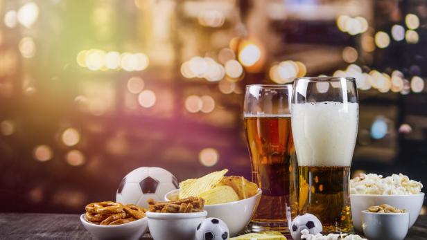 Zwei Gläser Bier, Snacks und Fußbälle auf einem Tisch vor verschwommenem Hintergrund.