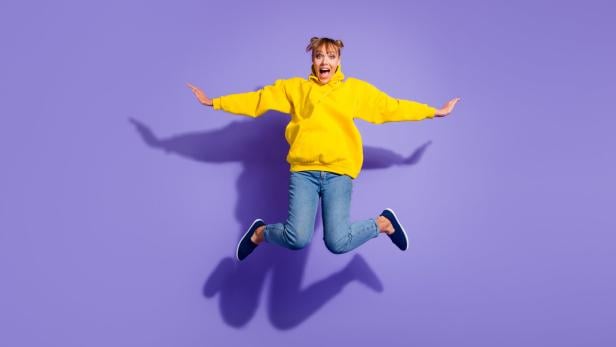 Junge Frau mit gelbem Hoodie springt fröhlich mit ausgebreiteten Armen vor lilafarbenem Hintergrund.