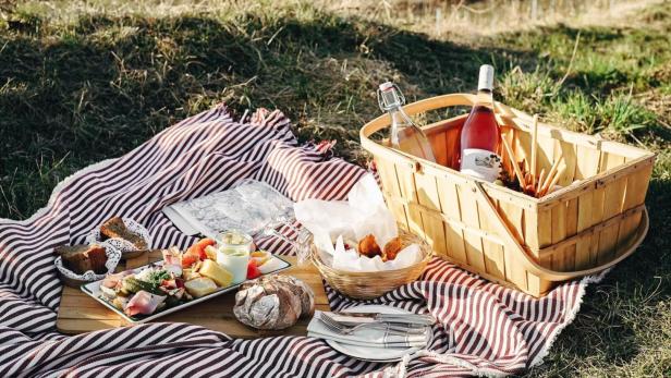 Ein Picknickkorb und Speisen auf einer rot-weiß gestreiften Decke im Grünen.
