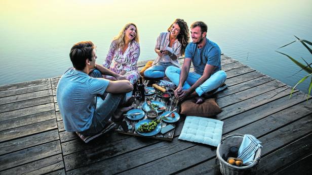 Eine Gruppe Freunde genießt ein Picknick auf einem Holzsteg am See.