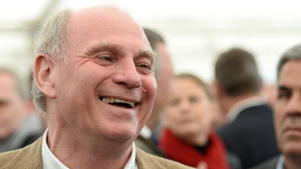 Ein lächelnder Mann mit hellem Haar und brauner Jacke im Gespräch mit anderen Personen.