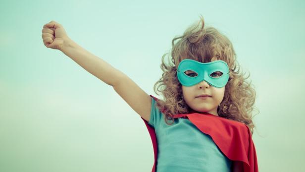 Ein Kind mit Superheldenmaske und rotem Umhang reckt den Arm in die Höhe.