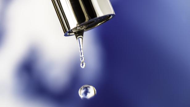 Ein Wassertropfen fällt aus einem silbernen Wasserhahn vor unscharfem blau-weißem Hintergrund.