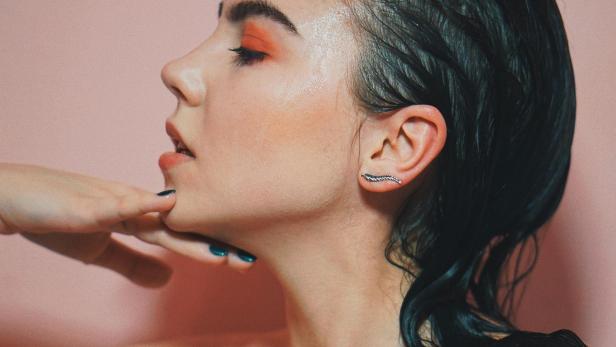 Eine Frau mit nassen, dunklen Haaren und orangefarbenem Lidschatten posiert vor einem rosa Hintergrund.