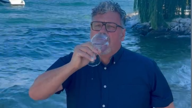 Ein Mann mit Brille trinkt ein Glas Wasser vor einer Küstenlandschaft.