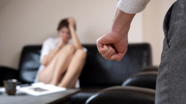 Eine Frau sitzt verängstigt auf einem Sofa, während eine geballte Faust bedrohlich im Vordergrund zu sehen ist.